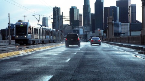 Dos autos van en dirección oeste por el Puente de la Primera, al lado de un tren de la Línea Dorada en dirección este.