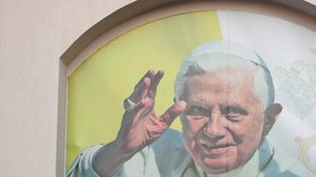 Varias personas se sientan delante de una pancarta colocada sobre una pared con una imagen del Papa Benedicto XVI que dice en español "León está listo para recibir al Papa y el mundo".