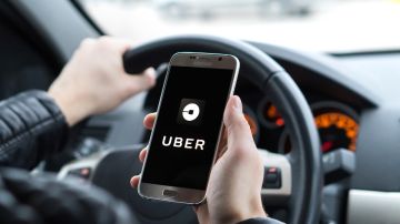 Una persona sostiene en una de sus manos un teléfono móvil con un logotipo de Uber en la pantalla, mientras conduce un auto.