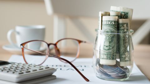 Una imagen con un pequeño vaso en el que hay billetes de dólar, unas gafas y una calculadora.