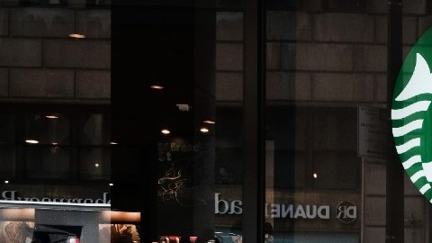 Una mujer camina frente a una sucursal de la cadena Starbucks, en la que se distingue su logotipo en color verde y blanco.