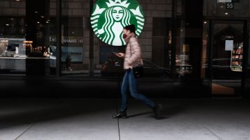 Una mujer camina frente a una sucursal de la cadena Starbucks, en la que se distingue su logotipo en color verde y blanco.