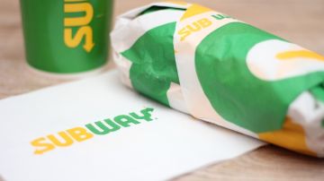 Un sandwich envuelto en papel de Subway con una servilleta de la misma marca y un vaso de refresco de la misma marca en colores verde y amarillo.