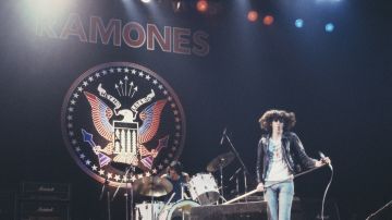 Joey Ramone (1951-2001), cantante de la banda de punk estadounidense The Ramones, en el escenario durante un concierto en vivo de la banda, con el baterista Tommy Ramone al fondo detrás de su batería, 1977.