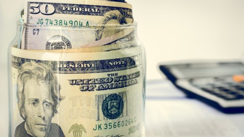 Imagen de un frasco en el que se guardan billetes, con una calculadora en el fondo.