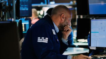 Una persona en el piso de ofertas en Wall Street se lleva la mano al rostro mientras mira pantallas con indicadores.
