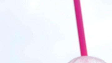 Imagen de dos envases de slurpee con popotes de colores sobre una superficie y en el fondo un logotipo de 7-Eleven.