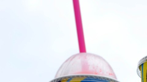 Imagen de dos envases de slurpee con popotes de colores sobre una superficie y en el fondo un logotipo de 7-Eleven.