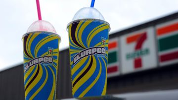 Imagen de dos envases de slurpee con popotes de colores sobre una superficie y en el fondo un logotipo de 7-Eleven.