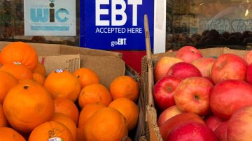 Un letrero en color azul indica que se aceptan tarjetas de comida EBT, el letrero está entre dos cajas con naranjas y manzanas.