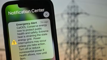 A photo illustration shows a background of electric power infrastructure with an Apple iPhone showing an Emergency Alert notification from CalOES urging the public to conserve energy to protect health and safety as the electricity grid is strained during a heat wave in Los Angeles, California on September 6, 2022. (Photo by Patrick T. FALLON / AFP) (Photo by PATRICK T. FALLON/AFP via Getty Images)