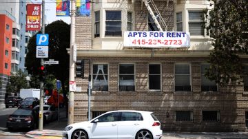 Imagen de un apartamento con las escaleras de emergencia visibles, un letrero con letras azules y rojas de alquiler y un auto blanco.