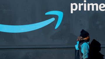 Un hombre vestido con un uniforme de Amazon color azul camina frente a un muro en el que se ve una sonrisa azul y letras blancas con la frase Prime.