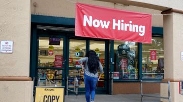 Una mujer entra a una tienda en donde cuelga una lona de color rojo en la que se anuncia contratación.