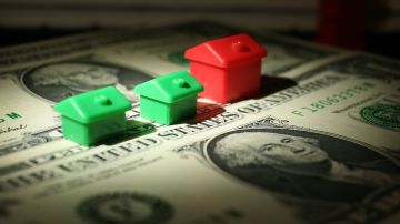 Imagen de dos casas de plástico de color verde junto a otra de color rojo, sobre billetes de dólares.