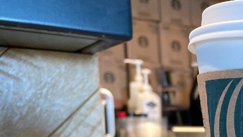 Imagen de un vaso con café de Starbucks, sobre una mesa de madera y en el fondo una persona que prepara café.