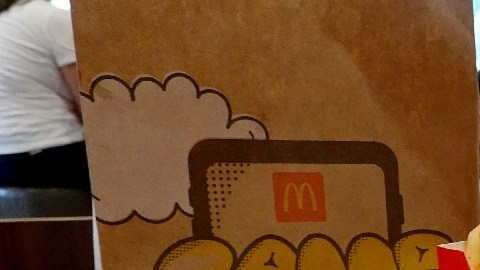 Imagen de un paquete de comida de McDonalds con una bolsa de papel, unas papas, un vaso con refresco y una hamburguesa.