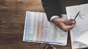 Imagen de una mesa con varios artículos de oficina y manos que están trabajando con papeles y computadoras.