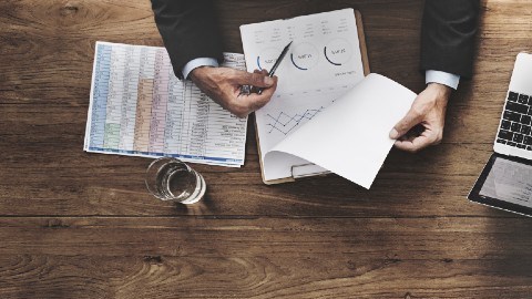 Imagen de una mesa con varios artículos de oficina y manos que están trabajando con papeles y computadoras.