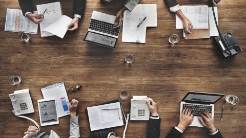 Imagen de una mesa con varios artículos de oficina y manos que están trabajando con papeles y computadoras.
