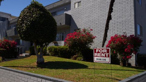 Imagen de un letrero de alquiler colocado en un jardín, frente a un complejo de apartamentos.