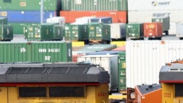 Imagen de vías de ferrocarril con una locomotora de color amarillo en el fondo y una bandera de Estados Unidos, además de un vehículo verde.