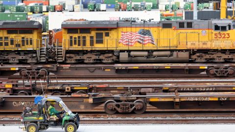 Imagen de vías de ferrocarril con una locomotora de color amarillo en el fondo y una bandera de Estados Unidos, además de un vehículo verde.