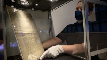 Una copia de la Constitución de los Estados Unidos se muestra detrás de una vitrina, mientras una persona extiende sus manos para sostenerla.