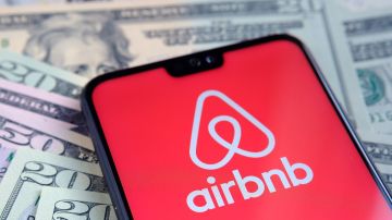 Imagen de un celular que está sobre unos billetes de color verde y en su pantalla de ve el logotipo de la empresa Airbnb en blanco con rojo.