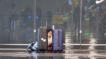 Imagen de una persona que está sentada en el piso de un aeropuerto en medio de dos maletas.