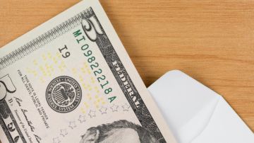 Imagen de un billete de cinco dólares dentro de un sobre de color blanco, sobre una mesa de madera.
