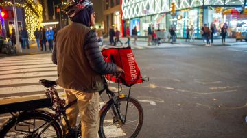 Imagen de una persona sobre una bicicleta, en medio de una calle, mientras lleva una bolsa de color rojo de reparto.
