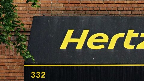 Imagen de un local con una marquesina de color negro con letras amarilla de la marca Hertz, y un vehículo blanco estacionado.