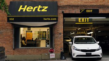 Imagen de un local con una marquesina de color negro con letras amarilla de la marca Hertz, y un vehículo blanco estacionado.