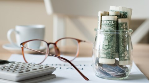 Imagen de un vaso lleno de billetes, junto con una calculadora y unos lentes.