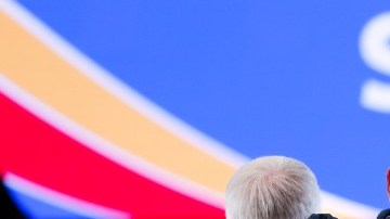 Imagen de dos personas que están sentadas en un aeropuerto, en medio de maletas de equipaje.