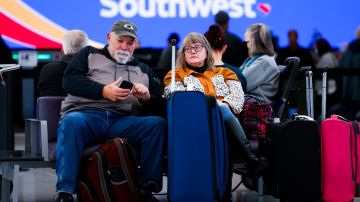 Imagen de dos personas que están sentadas en un aeropuerto, en medio de maletas de equipaje.