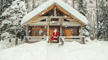 Imagen de una cabaña nevada y de un Santa Claus de pie frente a su puerta.