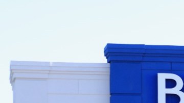 Imagen de una tienda de la marca Bed Bath & Beyond con la entrada en un marco de color azul y varios autos estacionados.