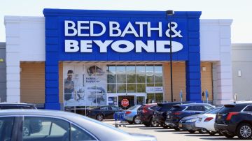 Imagen de una tienda de la marca Bed Bath & Beyond con la entrada en un marco de color azul y varios autos estacionados.