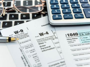Imagen de varios formularios para declarar impuestos, una pluma, una calculadora y el teclado de una computadora.