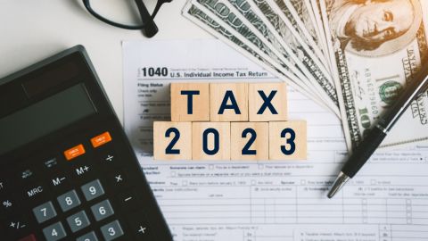 Imagen de varios billetes, una calculadora, unas gafas, una pluma y varios cubos de madera con la leyenda "tax 2023".