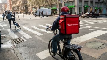 Imagen de una persona de reparto de DoorDash que va en una bicicleta en una calle entre edificios.