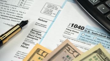 Imagen de varios formularios de impuestos, tres billetes, una calculadora y una pluma.