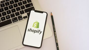 Imagen de la pantalla de un teléfono celular con el logotipo de la empresa Shopify, colocado sobre el teclado de una computadora.