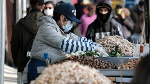 Imagen de una persona que selecciona frutas en un estante, en medio de la calle, mientras otras personas caminan.