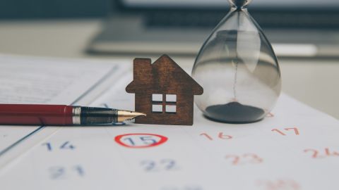 Imagen de una casa en miniatura de madera, sobre un calendario en el que está marcado el 15 con un círculo rojo y está junto a un reloj de arena y a una pluma.