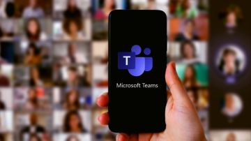 Imagen de una mano que sostiene un teléfono celular en cuya pantalla se ve el logotipo del programa Microsoft Teams en color negro y morado.