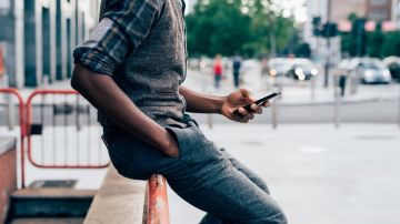 Imagen de un joven millenial sentado en el borde de una barda, mientras sostiene un teléfono celular.