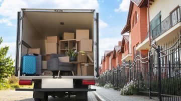 Imagen de un camión con varias cajas y artículos de hogar en la caja, estacionado frente a un conjunto de casas.